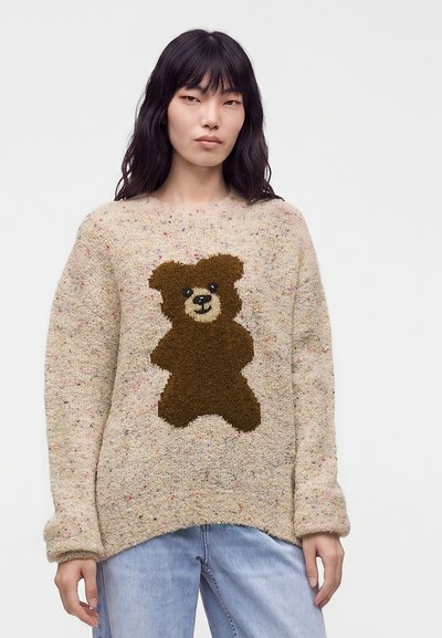 Personne portant un pull beige moucheté avec un grand motif d'ours en peluche marron, associé à un jean bleu clair.