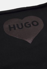 Tessuto nero con un grande design a forma di cuore metallico e la parola "HUGO" in carattere grassetto e scuro. Texture morbida, vista ravvicinata.