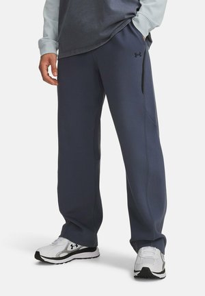 Man wearing dark blue Under Armour sweatpants and white Under Armour sneakers standing with one hand in pocket.