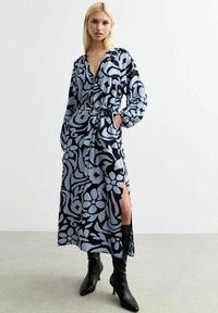Langes Blumenmuster-Kleid in Hellblau und Schwarz, mit V-Ausschnitt, langen Ärmeln, Gürtel in der Taille und Seitenschlitzen, kombiniert mit schwarzen Stiefeln.