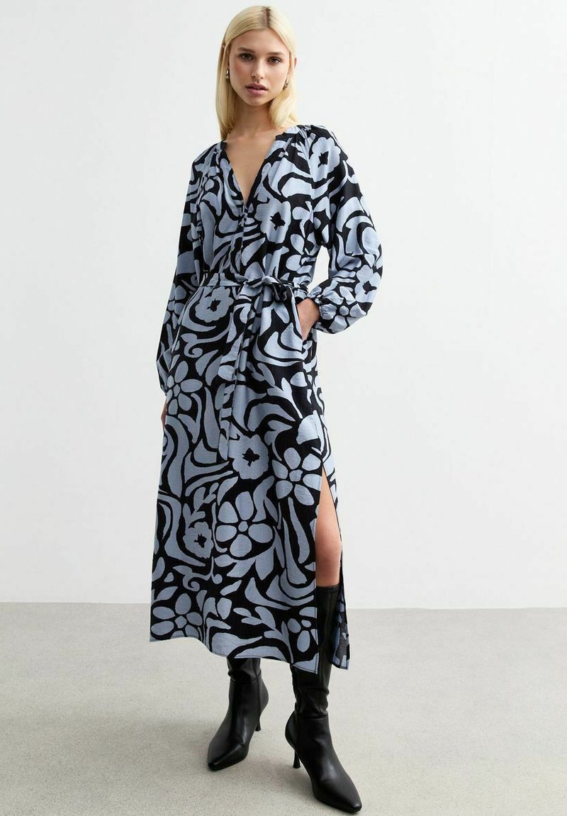 Langes Blumenmuster-Kleid in Hellblau und Schwarz, mit V-Ausschnitt, langen Ärmeln, Gürtel in der Taille und Seitenschlitzen, kombiniert mit schwarzen Stiefeln.