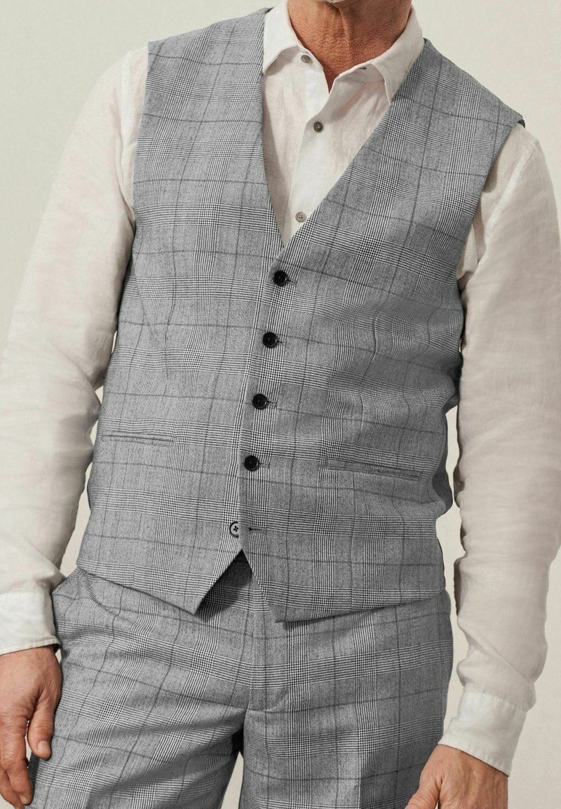 Gilet a quadri grigio realizzato in un tessuto testurizzato, con scollatura a V, sei bottoni neri e due tasche laterali, abbinato a una camicia bianca.