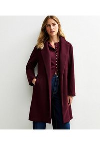 Cappotto in misto lana bordeaux con colletto a reverse, maniche lunghe e tasche frontali. Abbinato a un top con bottoni coordinato e jeans in denim.