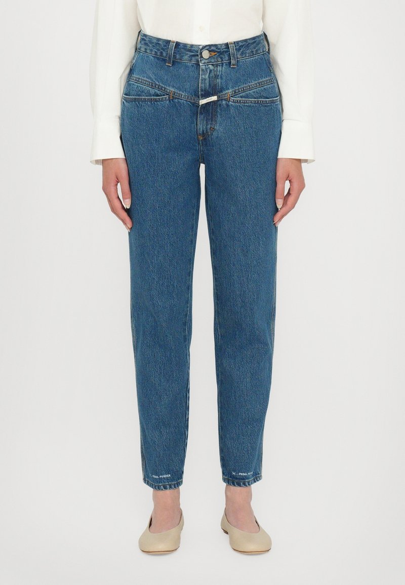 Personne portant un jean bleu taille haute avec des poches diagonales à l’avant, une chemise blanche à manches longues et des chaussures plates beiges, debout sur fond blanc.