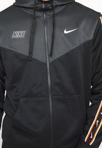 Svart Nike zip-up hoodie med grå panelering, har en kangaroo-ficka och orange-randiga detaljer på ärmarna. Inkluderar justerbar huva.