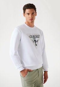 White sweatshirt with a triangular graphic design featuring the text "GUESS" in black, accented with green and silver details.