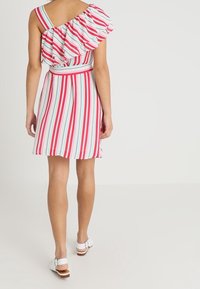 Monivärinen raidallinen mekko, jossa on yksi olka ja röyhelömuotoilu, sisältäen vaaleanpunaisia, punaisia ja sinisiä viivoja. Mukana on sopiva vyö ja valkoiset sandaalit.