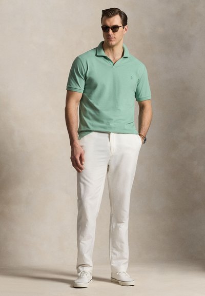 Άνδρας με πράσινη polo μπλούζα με κοντά μανίκια, λευκό παντελόνι, λευκά αθλητικά παπούτσια, γυαλιά ηλίου και ρολόι χειρός, στέκεται μπροστά σε ουδέτερο φόντο.