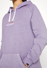 Personne portant un sweat à capuche ellesse violet avec des cordons blancs et une poche avant, affichant des logos sur la poitrine et le bord de la manche.