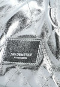 Pelle sintetica silver trapuntata con una texture liscia e lucida. Presenta un'etichetta nera rettangolare impresso con "Seidenfelt" e cuciture bianche.