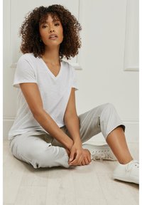Camiseta blanca de cuello en V hecha de un tejido suave, combinada con pantalones grises claros ajustados y zapatillas blancas, sentada en un suelo de madera clara.