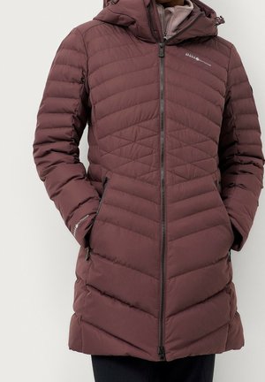 Persona con una chaqueta de invierno larga acolchada de color granate, con capucha, cremallera frontal y bolsillos laterales con cremallera, con las manos en los bolsillos.