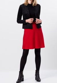 Blazer en suédine noire sur un col roulé noir, associé à une jupe évasée rouge et des bottines noires. Texture lisse, couleurs unies et design ajusté.