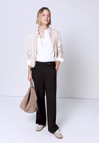 Femme aux cheveux courts portant une veste beige, une chemise blanche, un pantalon noir, des chaussures beiges, tenant un sac beige les yeux fermés sur un fond uni.