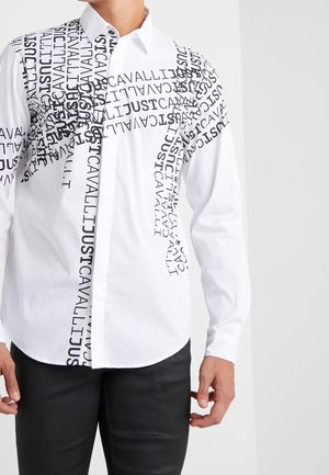 Hvid skjorte med knapper og sort spredt tekstdesign med teksten "JUST CAVALLI", båret af en mand i sorte bukser.