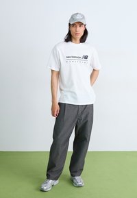 T-shirt en coton blanc avec le logo "new balance", pantalon baggy gris et baskets argentées. Porte aussi une casquette gris clair. Fond uni.