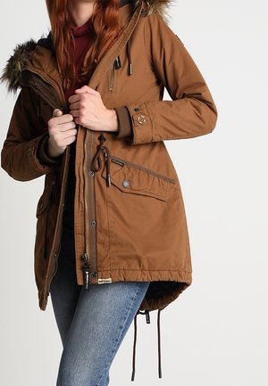 Kobieta w brązowej parkowej kurtce z kapturem wykończonym futerkiem, zapinająca zamek z przodu, ubrana w niebieskie dżinsy na jednolitym tle.