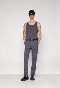 Szürke ujjatlan póló, slim fit szürke nadrág, fekete öv, fekete cipő, fekete sapka és napszemüveg. A tetoválásokkal ellátott modell egy sima háttér előtt áll.