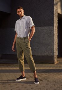 Camisa de mangas curtas às riscas em azul claro e branco, calças verde-azeitona e sapatos slip-on azul-marinho com solas brancas, de pé sobre um pavimento texturizado.