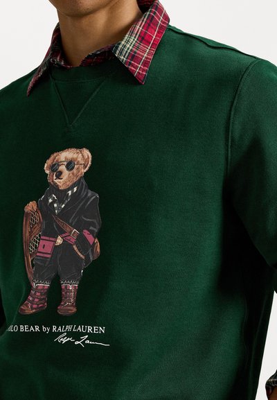 Zaļā džemperī ir liels grafiskais attēls ar lāci saulessargiem. Zem tā balti ir izdrukāts "POLO BEAR by RALPH LAUREN". Redzams sarkanais plānais apkakle.