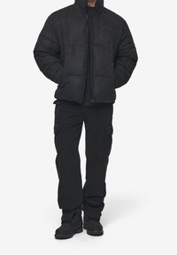 Persona che indossa una giacca imbottita nera, pantaloni cargo neri e stivali neri con lacci, in piedi con le mani nelle tasche della giacca su uno sfondo bianco.