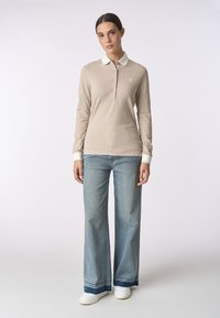 Polo beige a maniche lunghe con colletto bianco e placket a tre bottoni, abbinato a jeans a gamba larga blu chiaro e sneaker bianche.