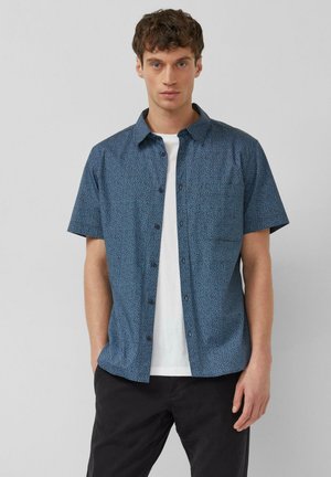 Giovane uomo che indossa una camicia blu a maniche corte con motivo a bottoni sopra una maglietta bianca e pantaloni neri, in piedi davanti a uno sfondo grigio uniforme.