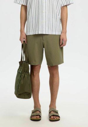 Shorts kaki avec un motif texturé, associés à une chemise courte à rayures boutonnée. Tenant un sac vert à cordon, portant des sandales grises.