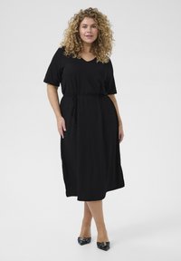Black V-neck dress with short sleeves, cinched waist tie, and knee-length hem. Smooth texture, simple silhouette, complemented by black heels.