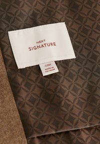 Tissu brun à motifs avec un design géométrique. Comprend une étiquette blanche avec "NEXT SIGNATURE" et des instructions d'entretien, cousue sur le tissu.