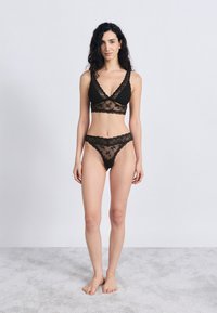 Conjunto de lingerie preta em renda, composto por um sutiã tipo bralette com decote em V e uma tanga combinando, apresentando padrões florais e detalhes em tecido transparente.