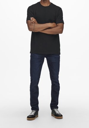 Man staat met armen gekruist, draagt een zwart T-shirt, donkerblauwe spijkerbroek en zwarte sneakers met witte veters en rubberkleurige zolen.