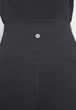 Black high-waisted leggings made of stretchy fabric featuring flatlock seams and a small white logo at the waist. Smooth texture and form-fitting design.