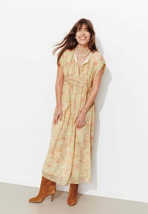 Femme souriante aux longs cheveux bruns, portant une robe midi jaune à fleurs, un collier de perles et des bottes à talons en daim marron, debout contre un mur blanc.