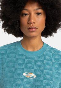 Tannengrüner Sweatshirt mit Struktur, das ein Gittermuster mit erhabenen Designelementen zeigt, sowie einem weißen Logo mit orangefarbenen Akzenten auf der Brust.