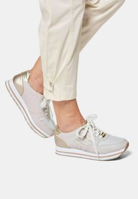Helle Sportschuhe, die eine Mischung aus glatten und strukturierten Materialien aufweisen, mit goldenen Akzenten und weißen Sohlen, gebunden mit Schnürsenkeln.