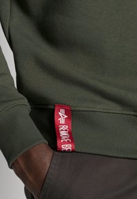 Maslinasto zeleni dukserica s rebrastim manžetama i crvenom etiketom na rubu s natpisom "REMOVE BE". Ugodna na dodir, tijelo prianjajući dizajn.