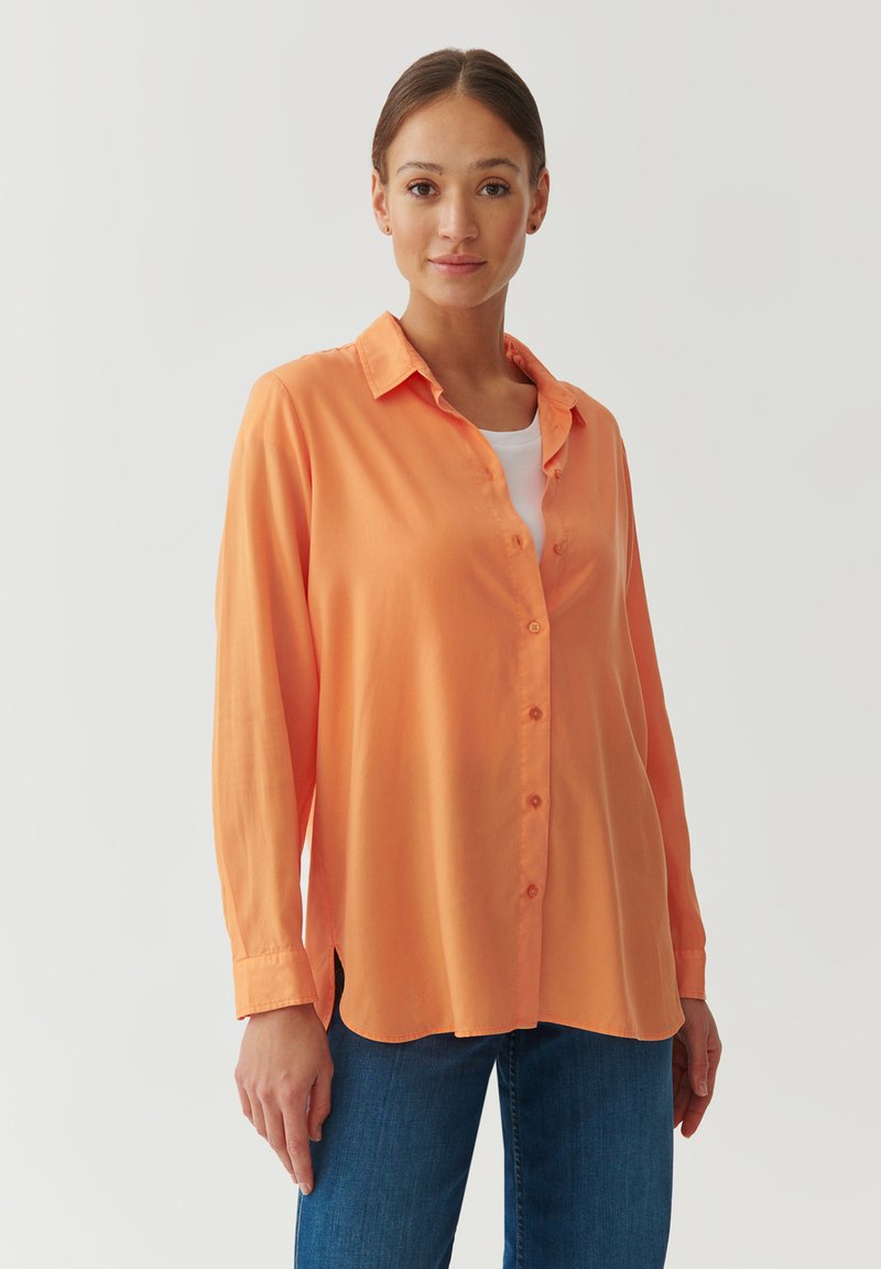Chemise boutonnée orange clair avec un col, des manches longues et une coupe décontractée. Portée par-dessus une chemise blanche, associée à un jean bleu.