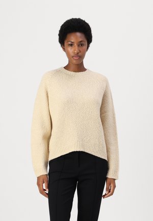 Beige strikkegenser med rund hals og senket skuldersøm. Har en teksturert overflate og en svakt forlenget bakhem.