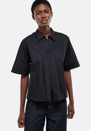 Femme portant une chemise noire à manches courtes avec un col et un pli à l'avant, ainsi qu'un jean bleu foncé, debout devant un fond blanc.
