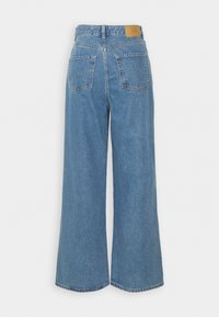 Plati mėlynos džinsinės kelnės su aukšta talija, dviem užpakalinėmis kišenėmis ir šviesiai rudos odos užsegimu gale. Minkšta, tekstūruota medžiaga.