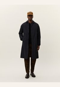 Marineblå lang frakk med knapper i front, brukt over en svart skjorte, sammen med mønstrede brune bukser og en lys caps med tekst.