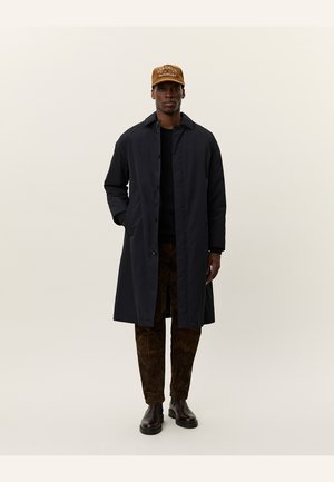Manteau long marine avec une fermeture à boutons, porté sur une chemise noire, associé à un pantalon marron à motifs et une casquette beige avec du texte.