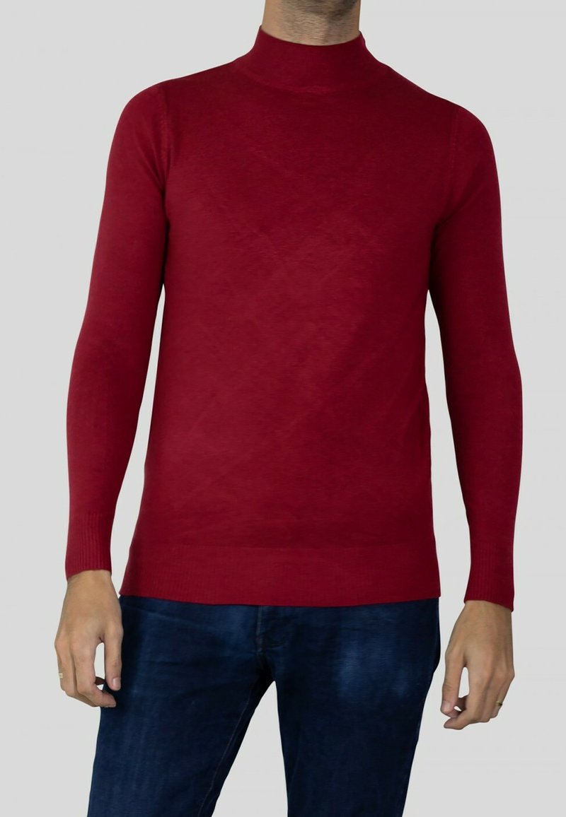 Homme portant un pull à col roulé rouge ajusté et un jean bleu foncé sur un fond gris uni.