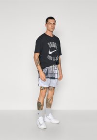 Sort oversized t-shirt med hvid tekst og Nike-logo; grå mønstrede shorts; hvide sneakers; synlige tatoveringer på arme og ben.