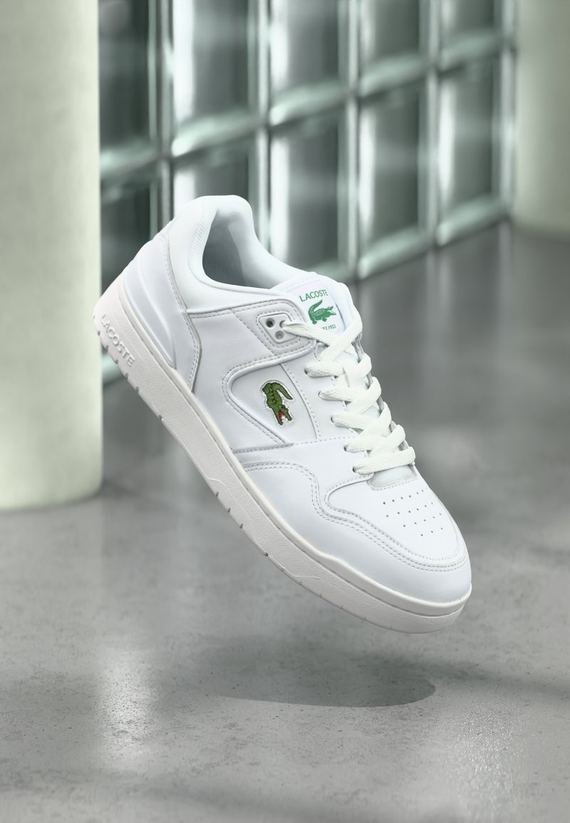 Άσπρο αθλητικό παπούτσι Lacoste με πράσινο λογότυπο κροκοδείλου στο πλάι και στη γλώσσα, αιωρούμενο πάνω από γκρι πάτωμα με φόντο τοίχο από γυάλινους κύβους.