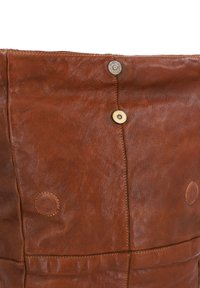 Brown leather bag with a textured surface, featuring two round brass accents, contrasting stitching, and a fold-over top design.