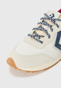 Baskets beiges avec une tige en mesh texturé, des accents bleu marine, des lacets à motifs et une semelle extérieure en caoutchouc brun avec un motif de semelle en zigzag.