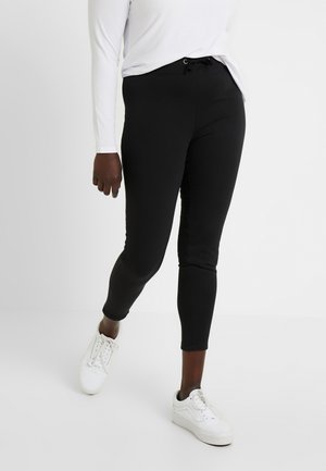 Black stretchy joggers with a drawstring waistband, featuring a slim fit and a smooth texture. Paired with white sneakers.