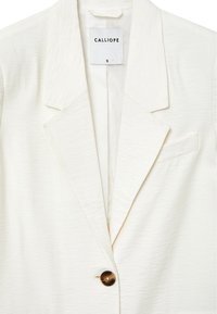 Blazer bianco realizzato in tessuto testurizzato, caratterizzato da un colletto a punta, chiusura con un singolo bottone e una discreta tasca sul petto. Etichetta del marchio visibile.